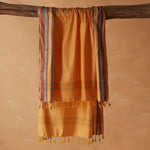 A yellow orange cotton pareo/sarong with multicolored striped borders and tassels, draped over a rustic wooden branch against a warm beige background.