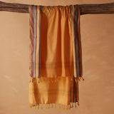 A yellow orange cotton pareo/sarong with multicolored striped borders and tassels, draped over a rustic wooden branch against a warm beige background.