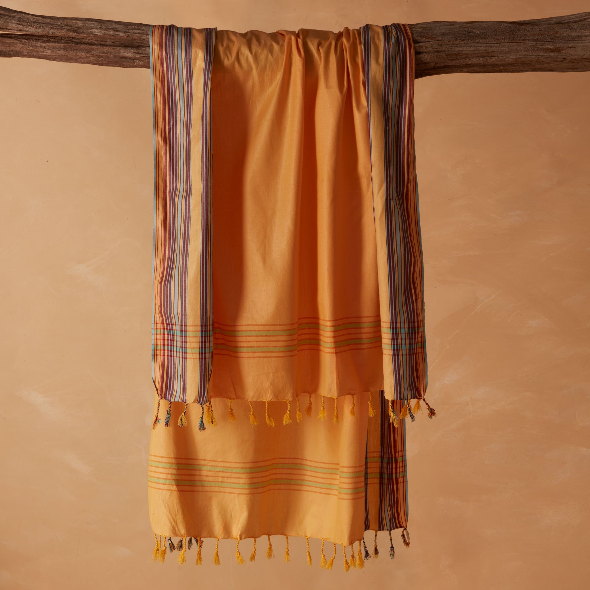 A yellow orange cotton pareo/sarong with multicolored striped borders and tassels, draped over a rustic wooden branch against a warm beige background.