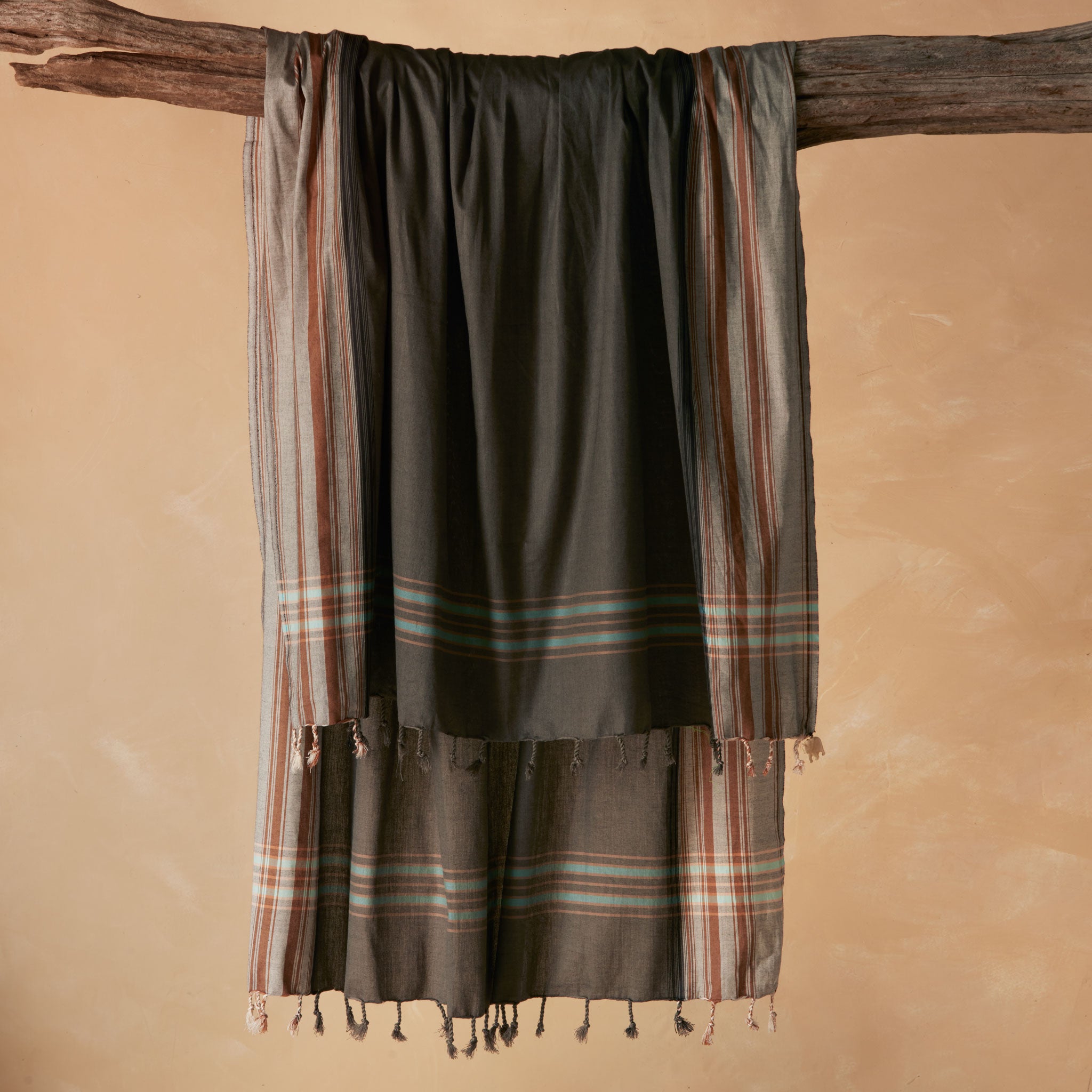 A dark gray cotton pareo/sarong with striped borders in beige, brown, and light blue, draped over a rustic wooden branch against a warm beige background.