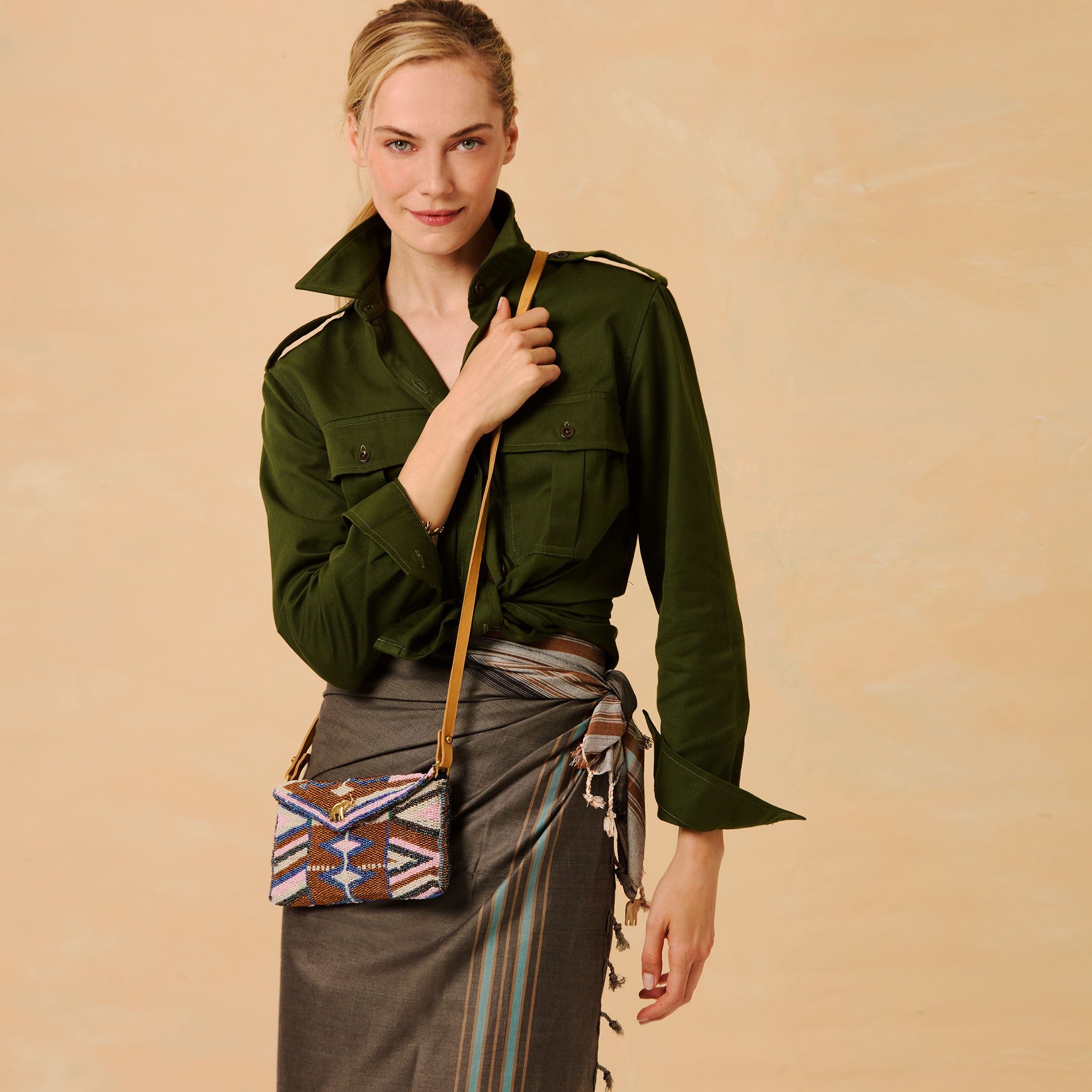 A woman models an olive green military-style shirt jacket with shoulder epaulets, paired with a dark grey pareo wrapped as a skirt and a small geometric-patterned crossbody bag with a tan strap, set against a solid beige background.