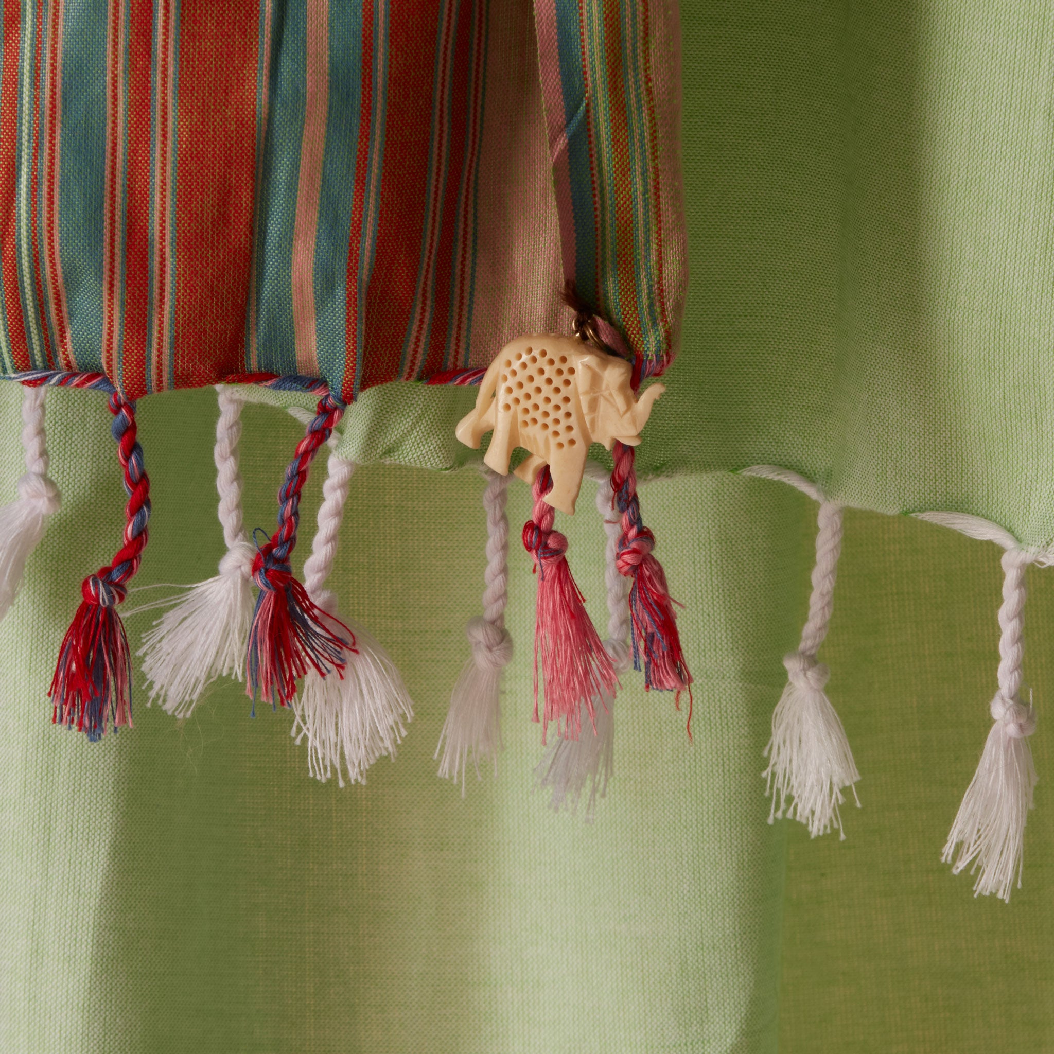 A close up of the tassels and a cream colored elephant charm of a light green woven pareo/sarong with red, blue and white stripes.