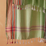 A light green cotton sarong with white tassels and multicolored striped borders, shown hanging against a solid warm beige colored background.