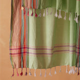 A light green cotton sarong with white tassels and multicolored striped borders, shown hanging against a solid warm beige colored background.