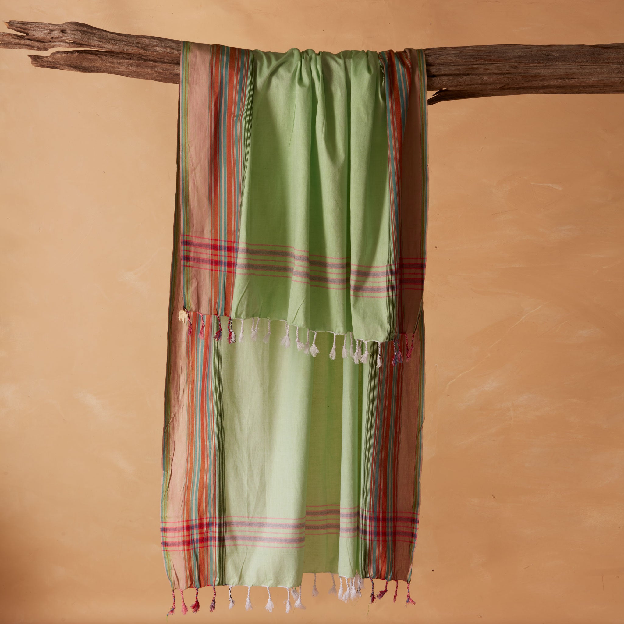 A light green woven pareo/sarong with red and white striped borders and white tassels, hanging from a rustic wooden branch against a warm beige background