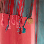 A close-up of a striped pareo/sarong with pink, red, and teal vertical bands, finished with braided tassels and a small beige elephant charm.