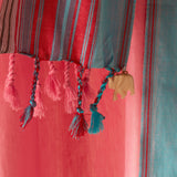 A close-up of a striped pareo/sarong with pink, red, and teal vertical bands, finished with braided tassels and a small beige elephant charm.