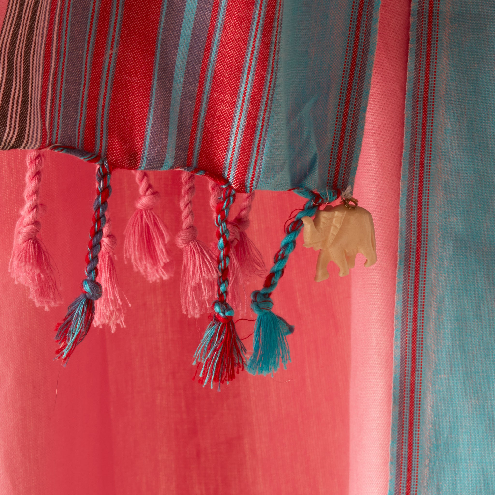 A close-up of a striped pareo/sarong with pink, red, and teal vertical bands, finished with braided tassels and a small beige elephant charm.