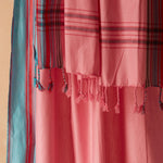 A close-up of a pink woven pareo/sarong with braided tassels and striped plaid sections in red, blue, black, and white, draped against a warm beige background.