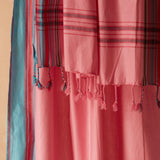 A close-up of a pink woven pareo/sarong with braided tassels and striped plaid sections in red, blue, black, and white, draped against a warm beige background.