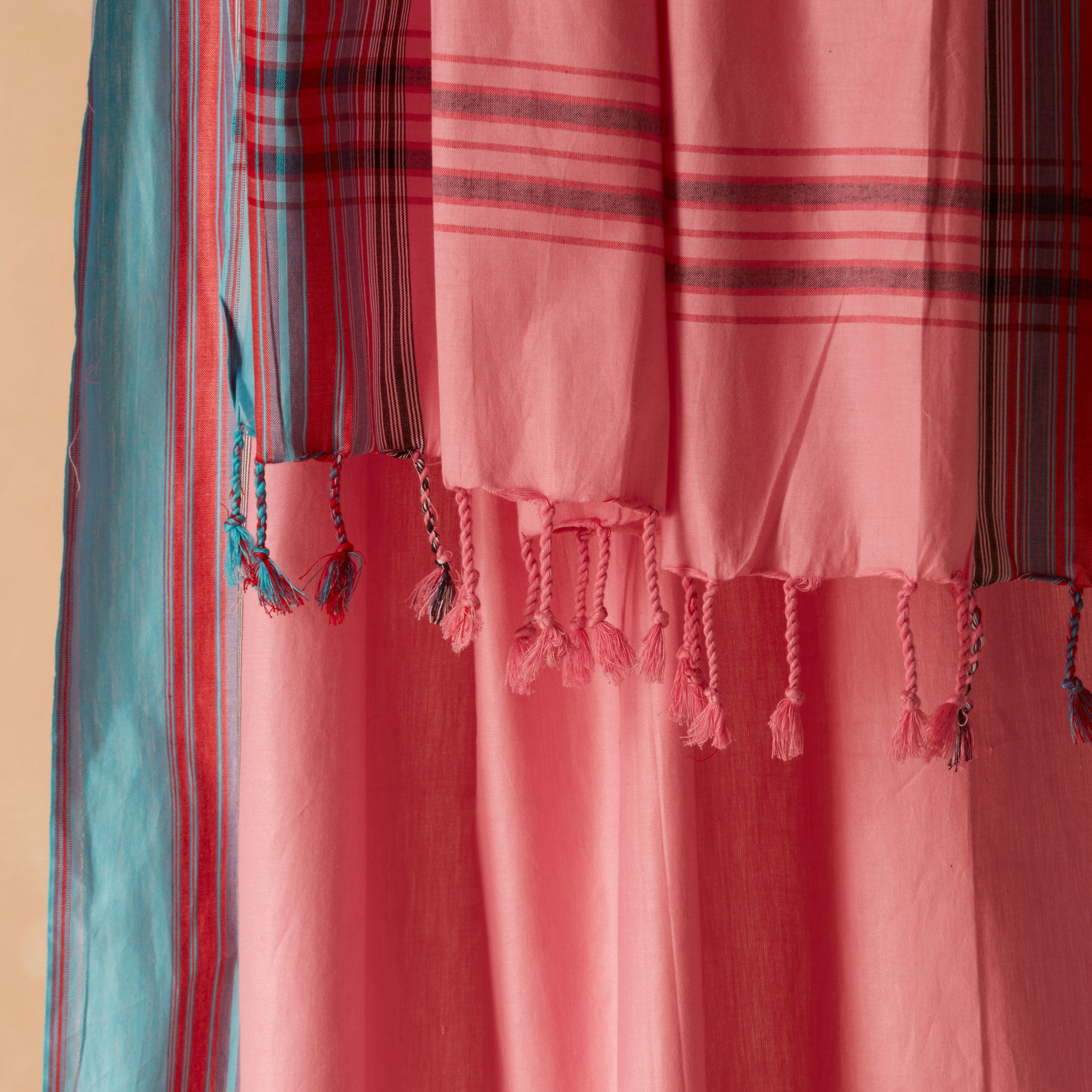 A close-up of a pink woven pareo/sarong with braided tassels and striped plaid sections in red, blue, black, and white, draped against a warm beige background.
