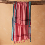 A pink and teal striped pareo/sarong with fringe tassels hanging from a rustic wooden branch against a warm beige background.