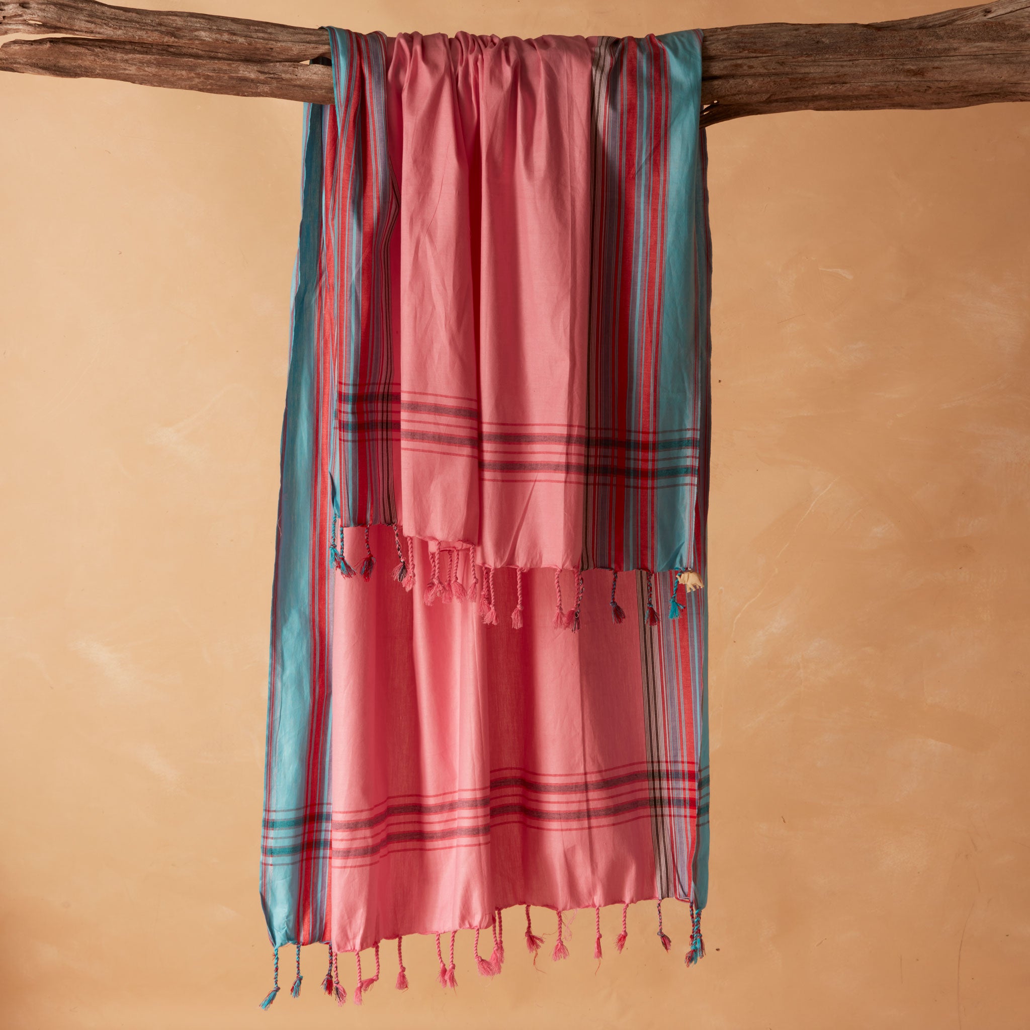 A pink and teal striped pareo/sarong with fringe tassels hanging from a rustic wooden branch against a warm beige background.