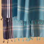 A detail view of a teal blue and navy striped cotton pareo/sarong with tassel fringe against a warm beige background.