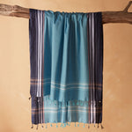 A teal blue and navy striped cotton pareo/sarong with tassel fringe, draped over a rustic wooden branch against a warm beige background.