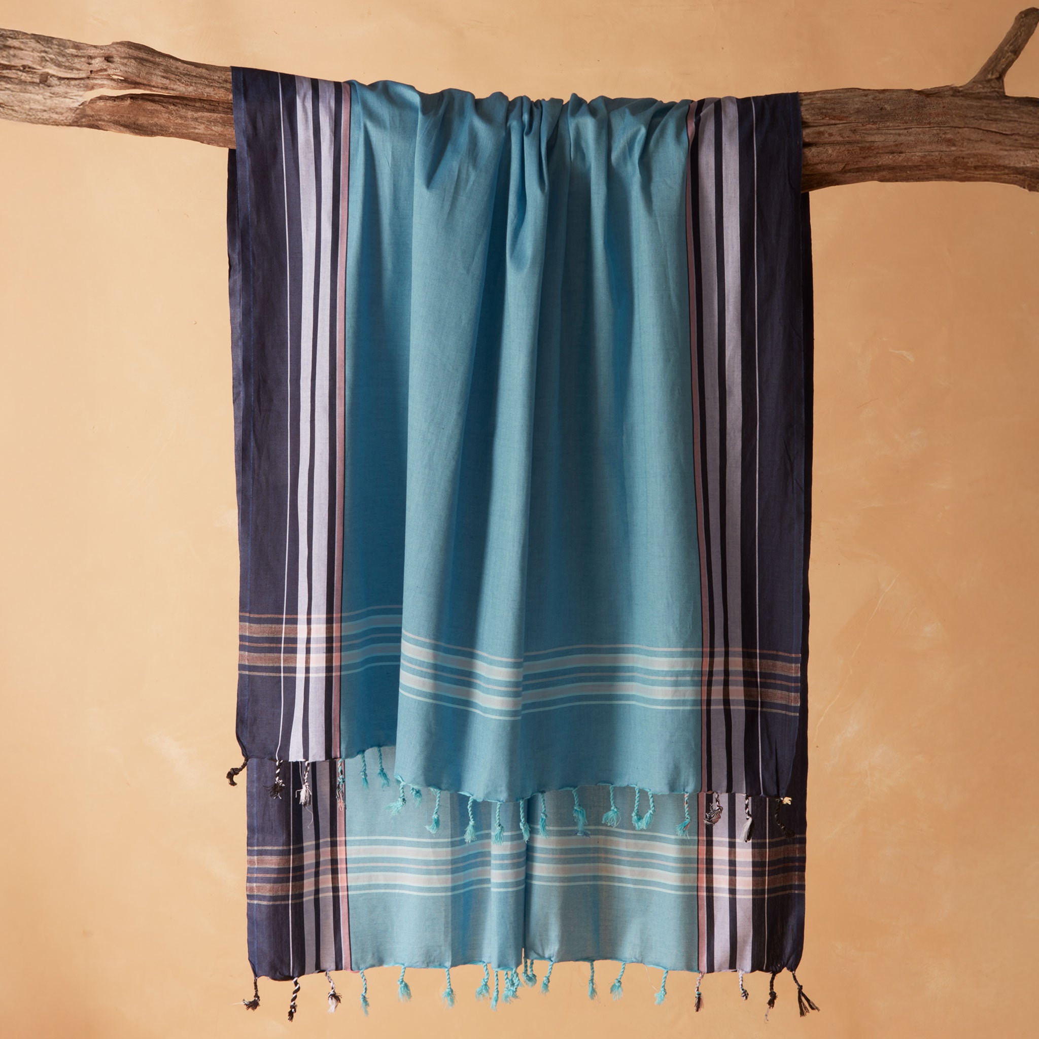 A teal blue and navy striped cotton pareo/sarong with tassel fringe, draped over a rustic wooden branch against a warm beige background.