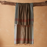 A draped pareo/sarong in grey and brown with red striped bands and tassel fringe, hanging from a rustic wooden branch against a warm beige background.
