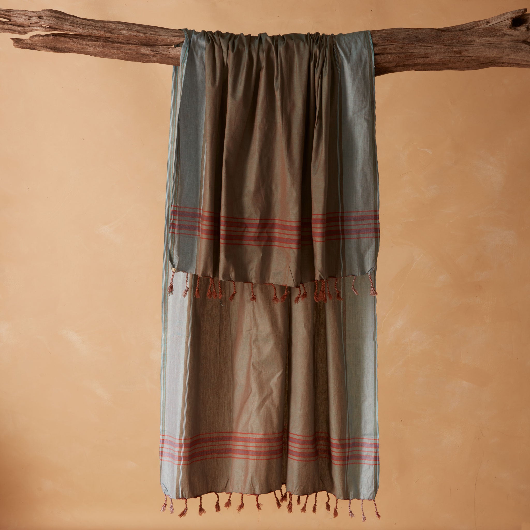 A draped pareo/sarong in grey and brown with red striped bands and tassel fringe, hanging from a rustic wooden branch against a warm beige background.