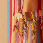 A close-up of a colorful striped pareo/sarong with braided tassels and a small cream elephant charm, shown against a warm beige background.