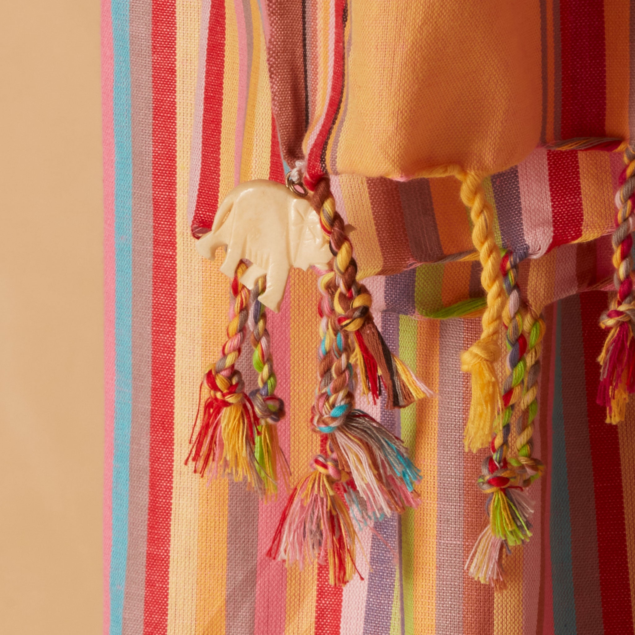 A close-up of a colorful striped pareo/sarong with braided tassels and a small cream elephant charm, shown against a warm beige background.