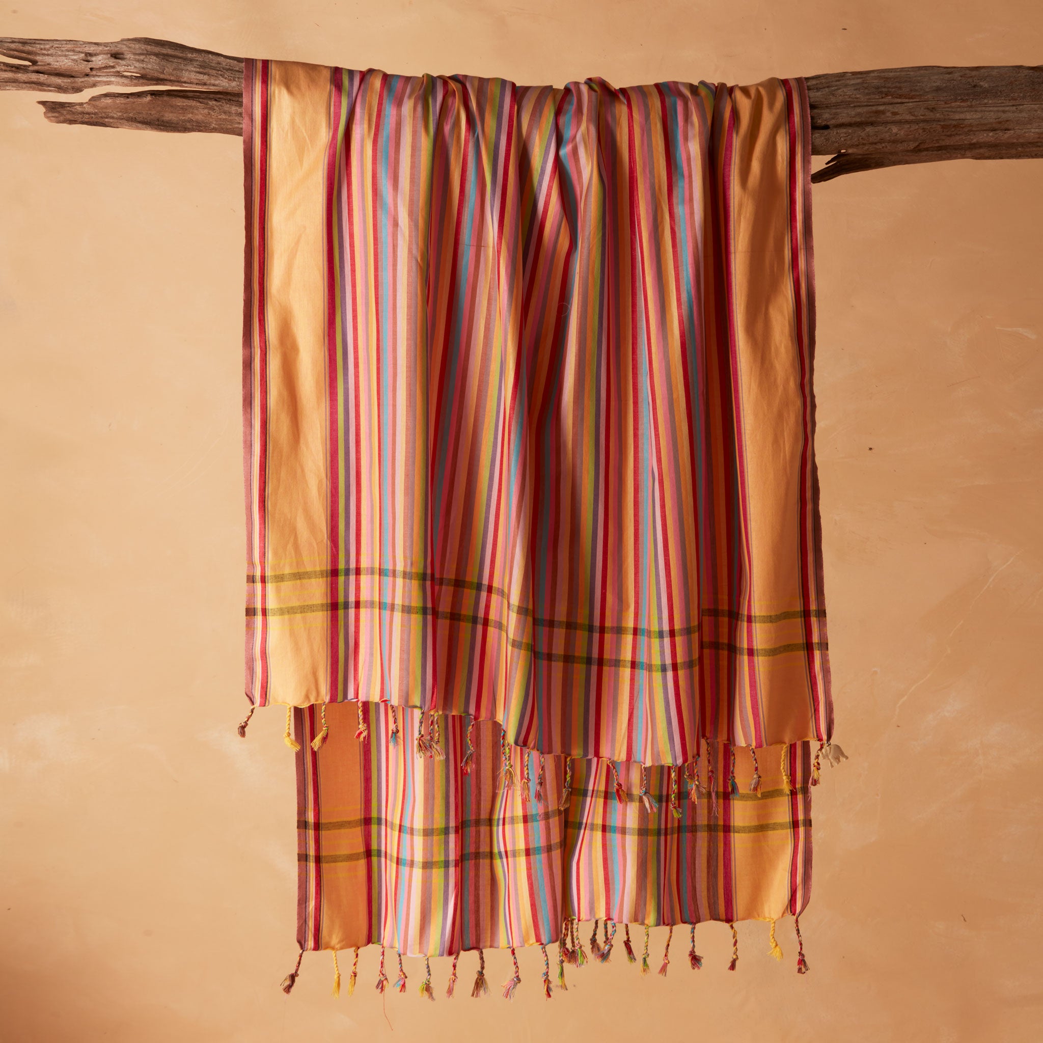 A multicolored striped pareo/sarong with fringed tassels draped over a rustic wooden branch against a warm beige background.