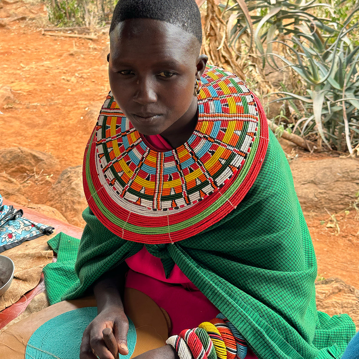 Woman wearing a colorful neck piece and green garment with stacked beaded bracelets sitting on the ground with natural foliage in the background