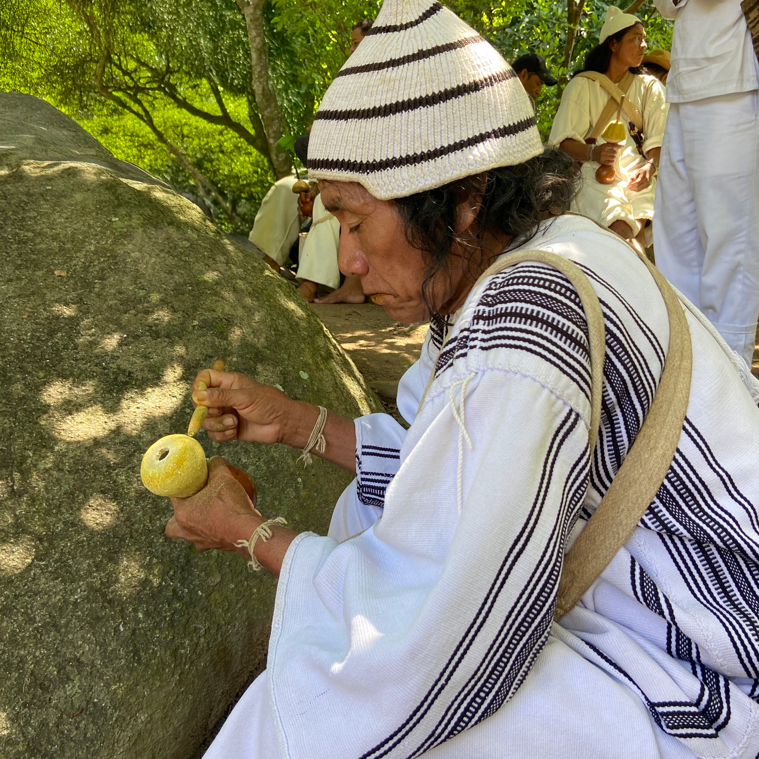 Person in traditional clothing with a white and black striped hat, sitting outdoors near a large rock.