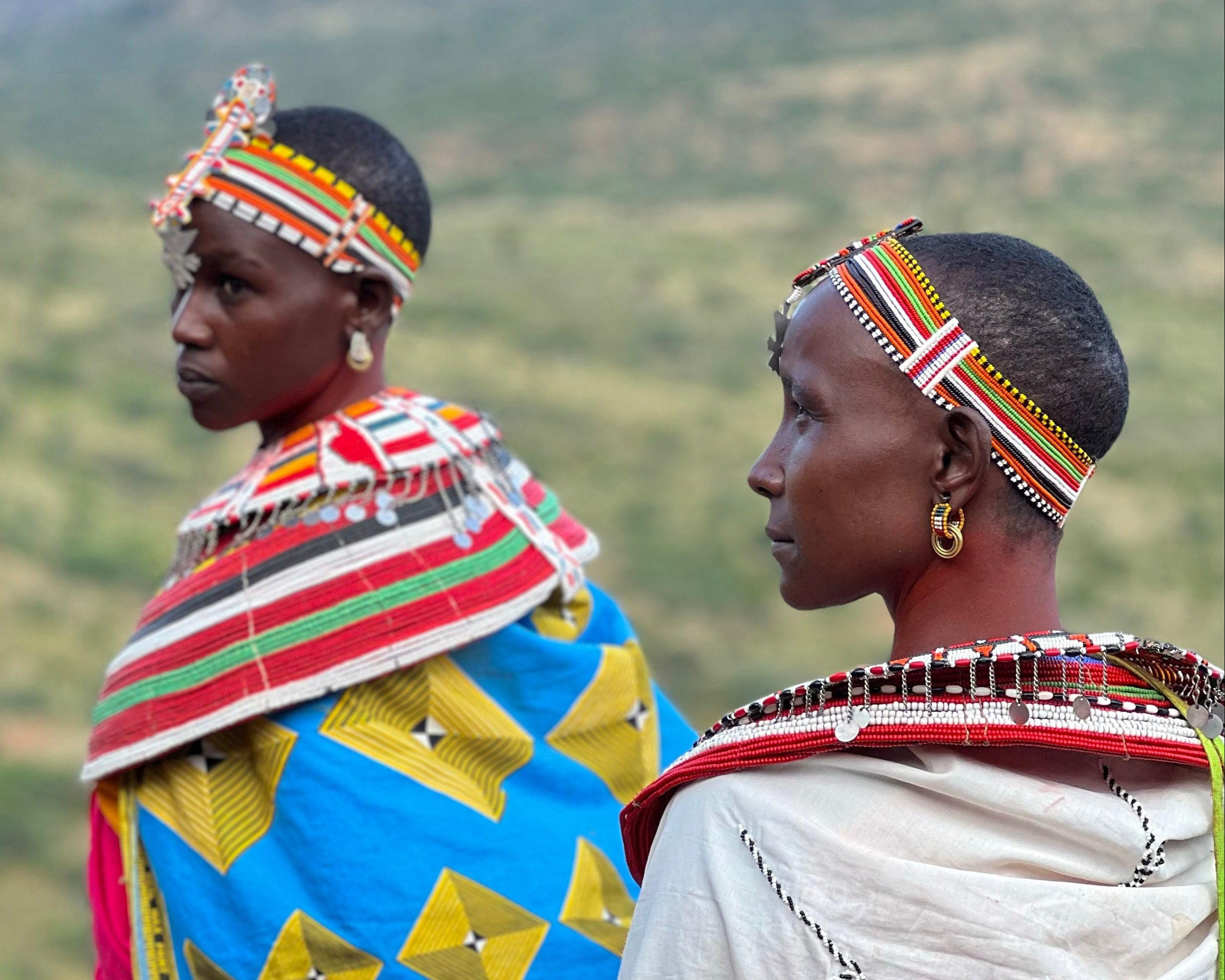 Two Kenayn women in traditional attire with colorful beaded headbands, earrings, and necklaces  with clothing clothing against a green landscape