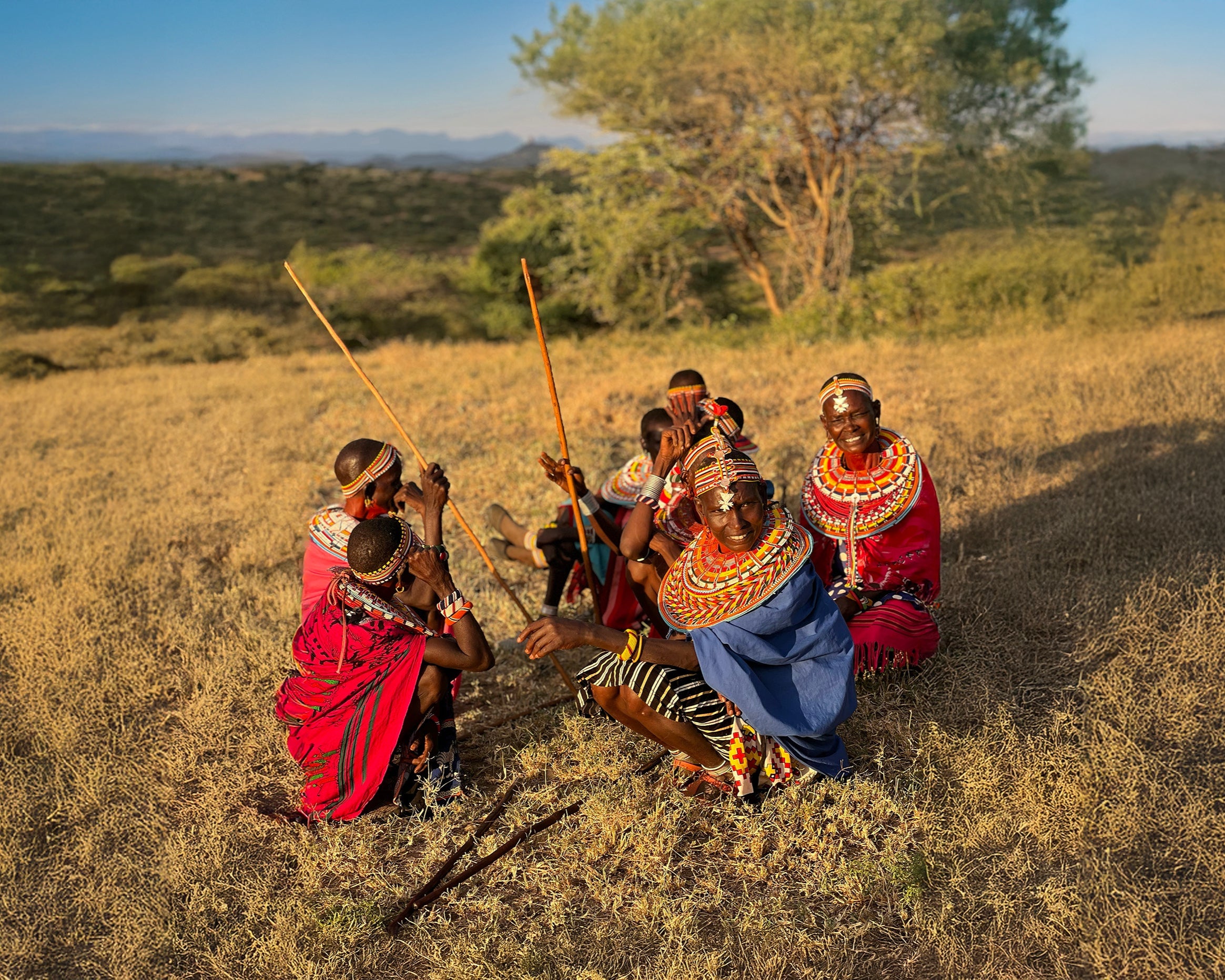 Group of Samburu people in traditional attire sitting in a small circle on a grassy field with trees in the background