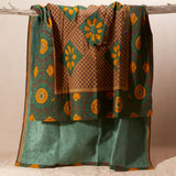 A draped patterned fabric in green, brown, and mustard yellow showing the towel solid green lining hanging from a rustic wooden branch against a neutral beige background.