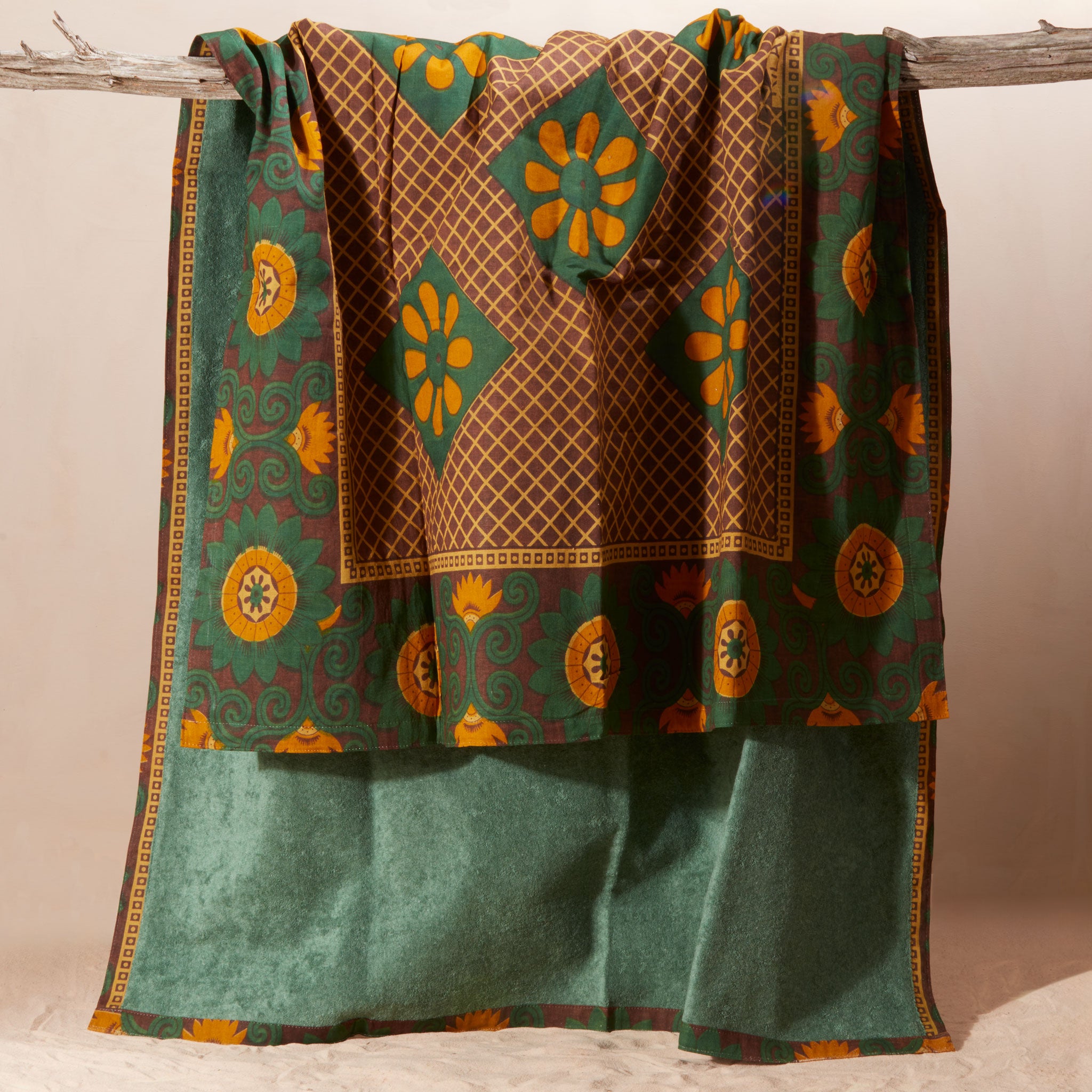 A draped patterned fabric in green, brown, and mustard yellow showing the towel solid green lining hanging from a rustic wooden branch against a neutral beige background.