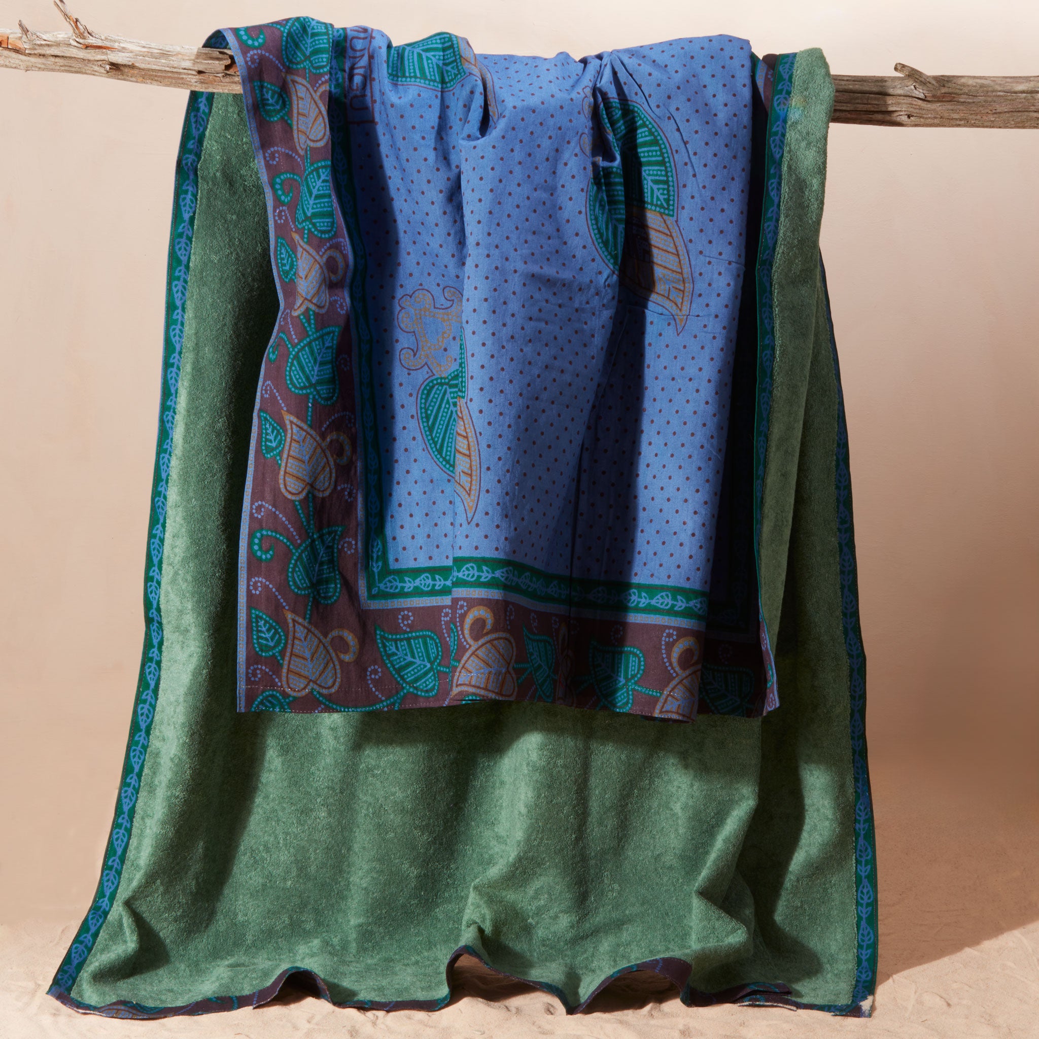 A Kanga towel featuring a blue printed cotton fabric with paisley and leaf motif featuring a green terry cloth lining hanging from a rustic wooden branch against a warm beige background.