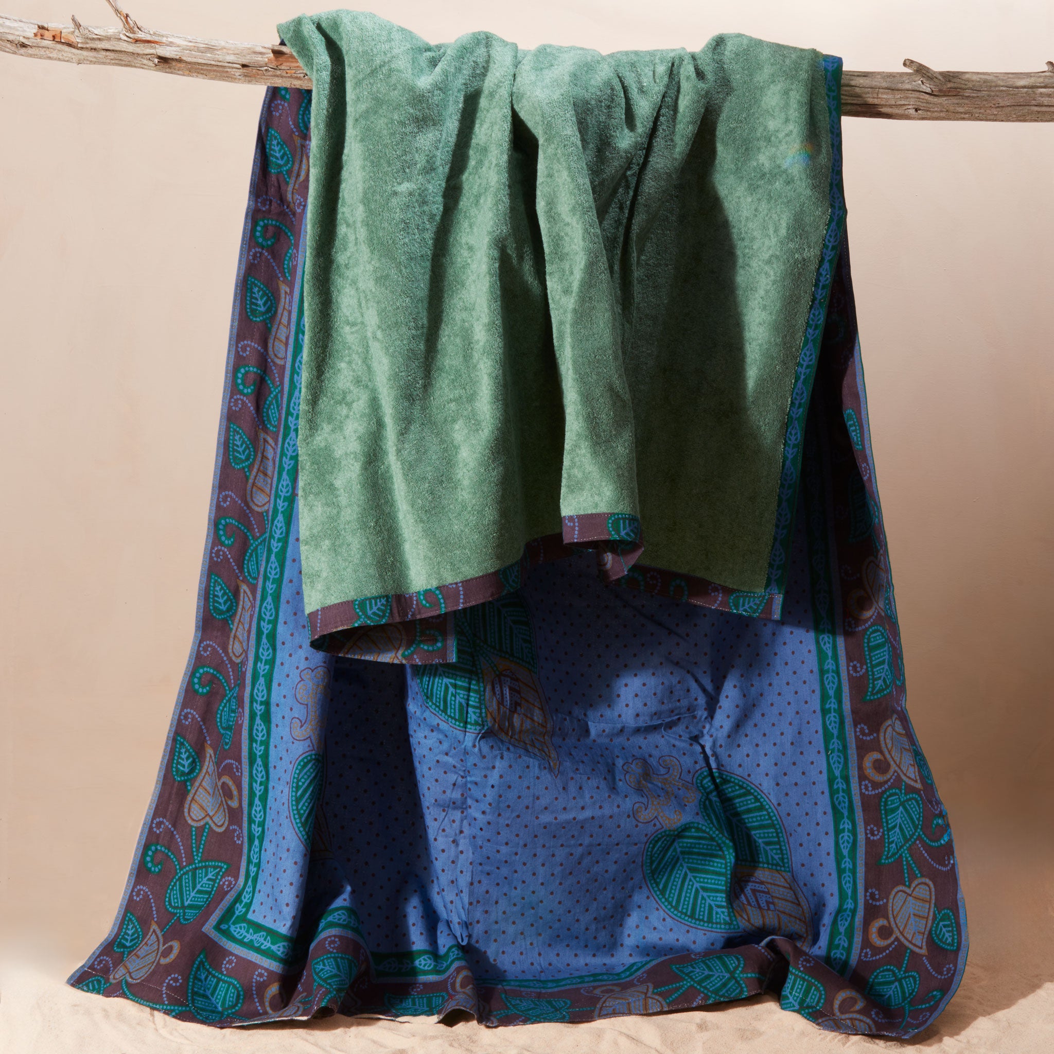 A folded and draped green towel hanging from a rustic wooden branch, with matching blue and teal patterned kanga fabric on the other side.