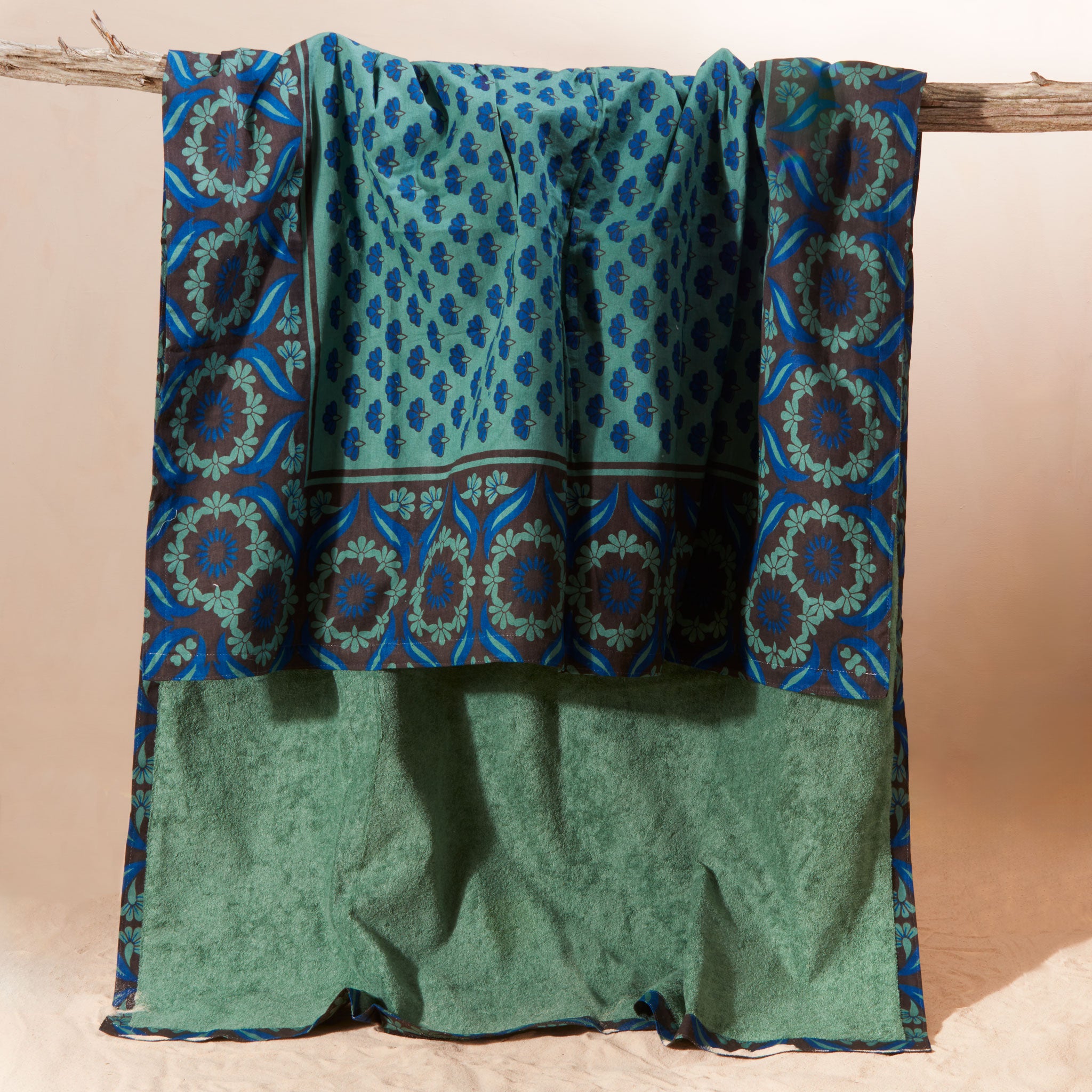 A blue and green towel  featuring a printed cotton fabric with geometric borders and a solid green terry cloth lining, draped over a rustic wooden branch against a beige background.