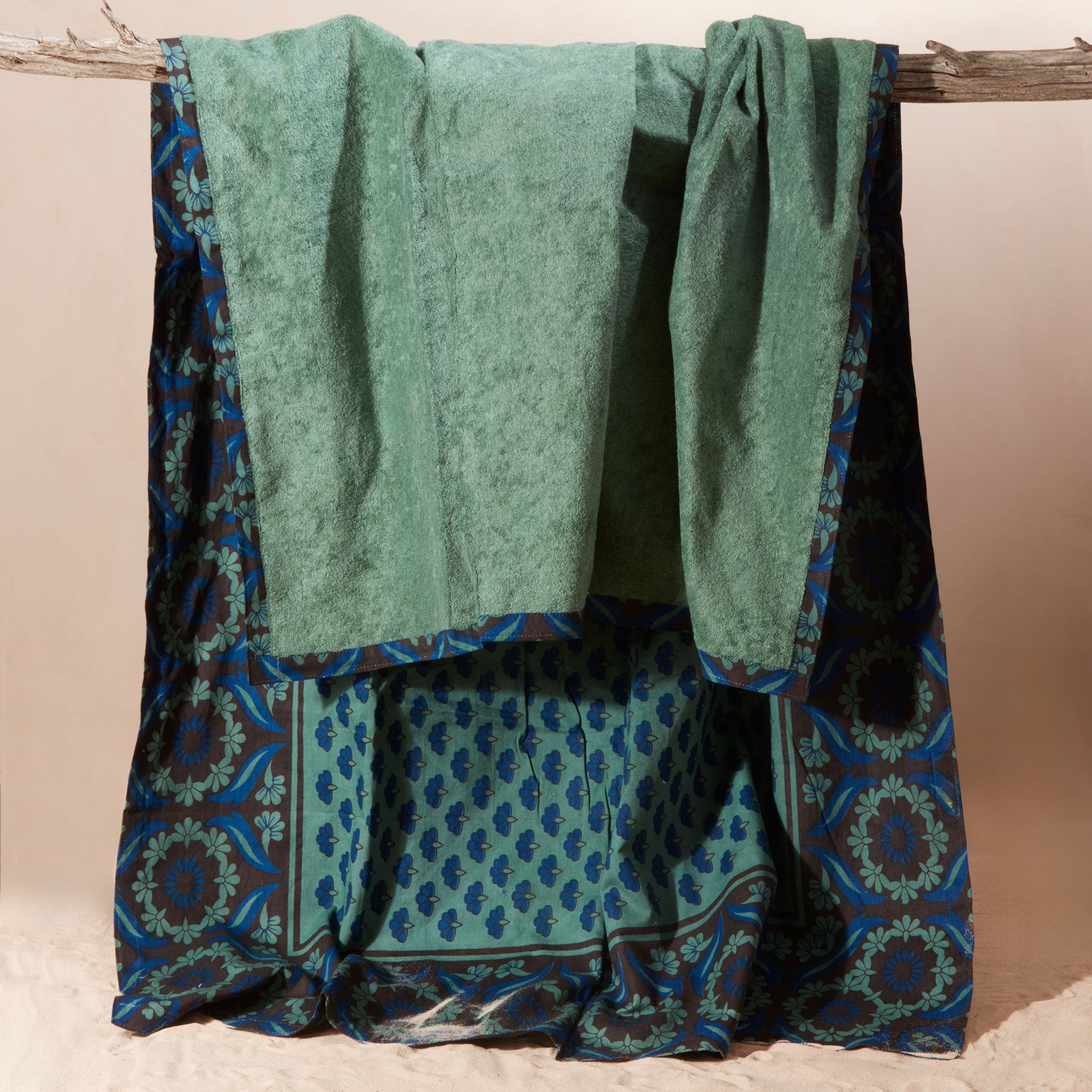 The solid green terry cloth of a blue and green towel  featuring a printed cotton fabric with geometric borders, draped over a rustic wooden branch against a beige background.
