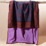 A draped piece of kanga fabric displayed on a rustic branch against a plain beige backdrop. The top section is dark blue with a repeating floral pattern in reddish-brown and border of reddish-brown with small blue accents, while the lower section showing the interior of the towel a solid light purple.