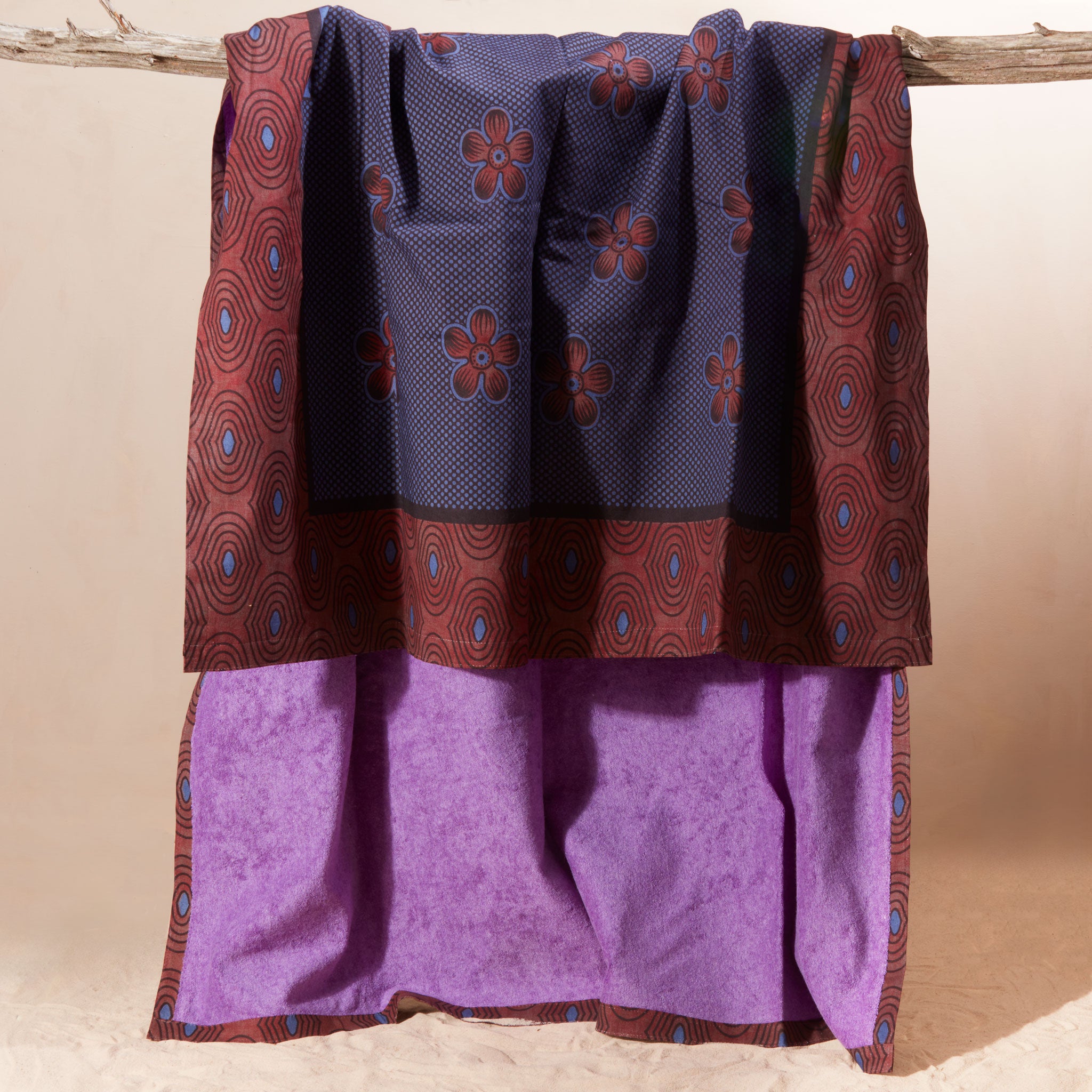 A draped piece of kanga fabric displayed on a rustic branch against a plain beige backdrop. The top section is dark blue with a repeating floral pattern in reddish-brown and border of reddish-brown with small blue accents, while the lower section showing the interior of the towel a solid light purple.
