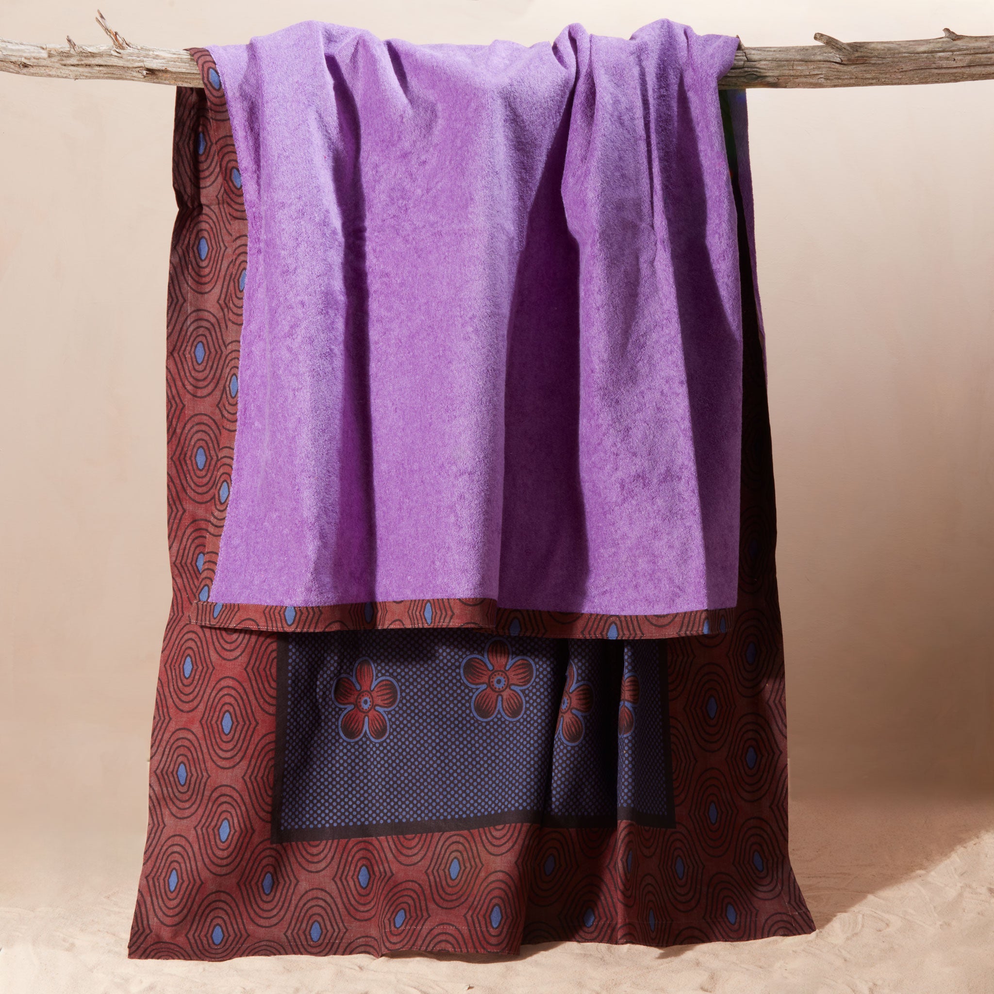 A purple towel draped over a rustic wooden branch showing the reddish brown, blue floral patterned side of the towel.