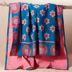 A kanga towel with a teal blue base, pink floral motifs, and magenta circular patterns on one side and pink terry cloth on the other side, hanging from a rustic wooden branch against a plain beige background.