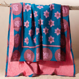 A kanga towel with a teal blue base, pink floral motifs, and magenta circular patterns on one side and pink terry cloth on the other side, hanging from a rustic wooden branch against a plain beige background.