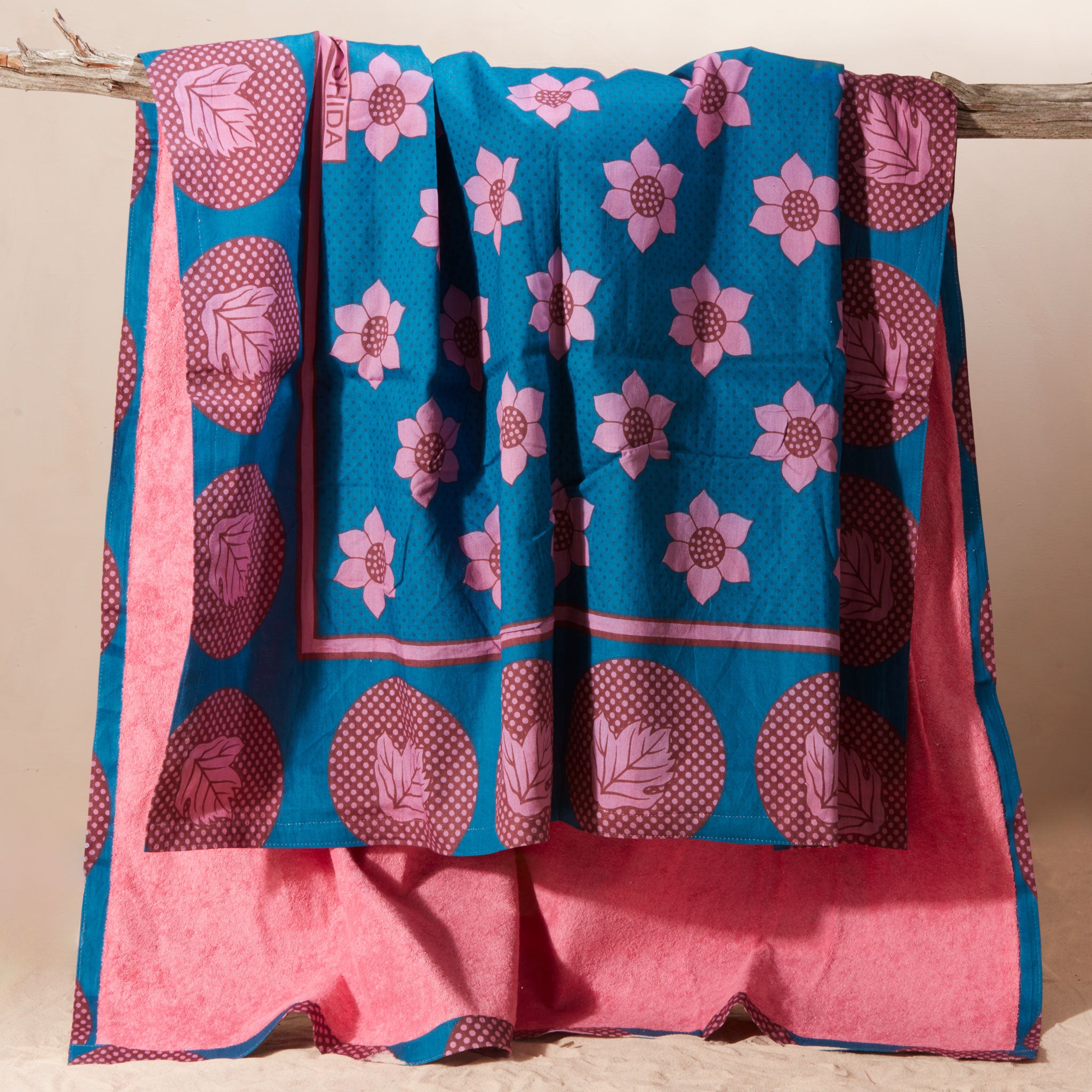 A kanga towel with a teal blue base, pink floral motifs, and magenta circular patterns on one side and pink terry cloth on the other side, hanging from a rustic wooden branch against a plain beige background.
