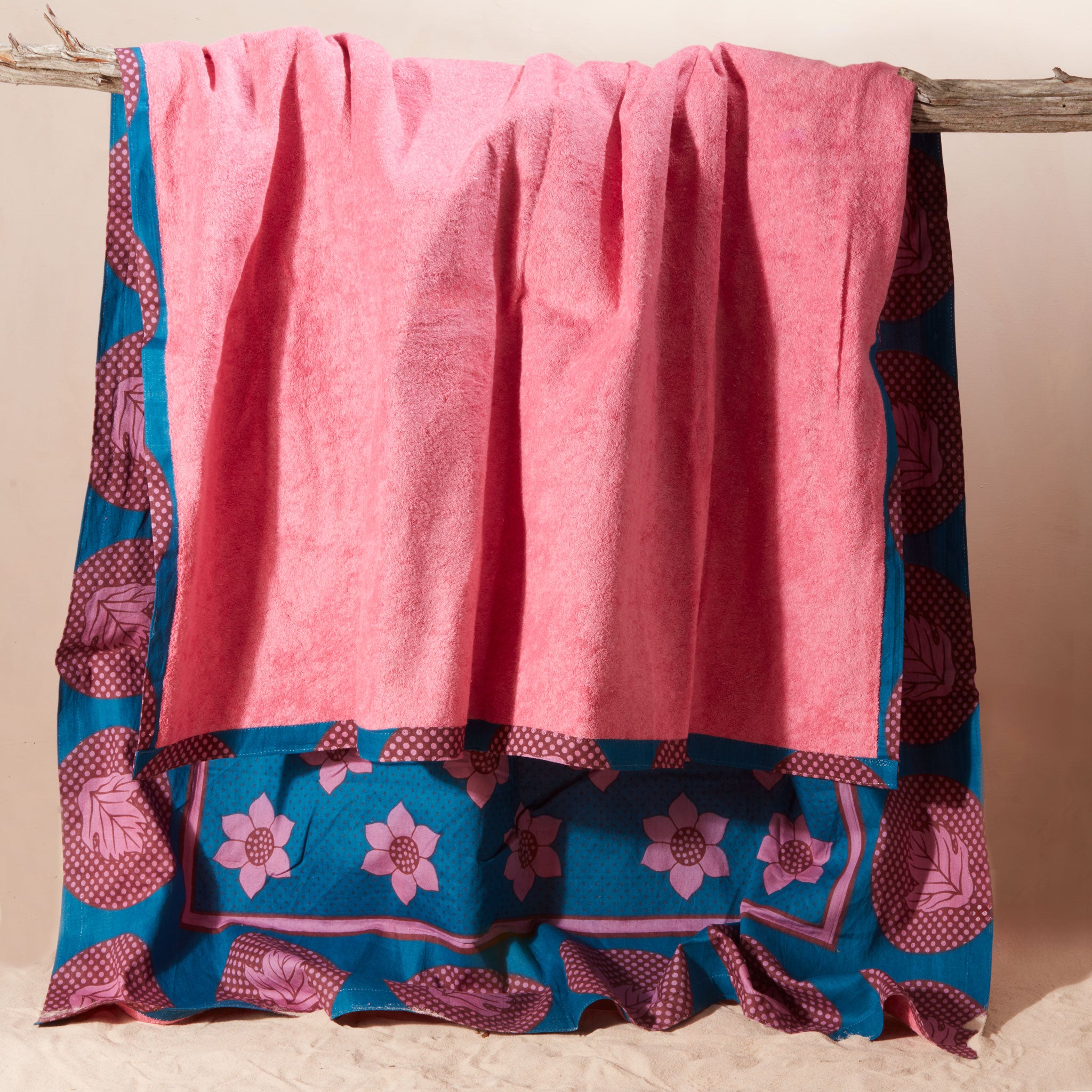 The pink terry cloth side of a kanga towel with a teal blue base, pink floral motifs, and magenta circular patterns, hanging from a rustic wooden branch against a plain beige background.