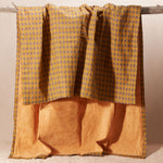 A draped kanga towel in warm mustard yellow and brown, featuring a patterned cotton side and a yellow-orange terry cloth side, hanging from a rustic wooden branch against a neutral beige background.