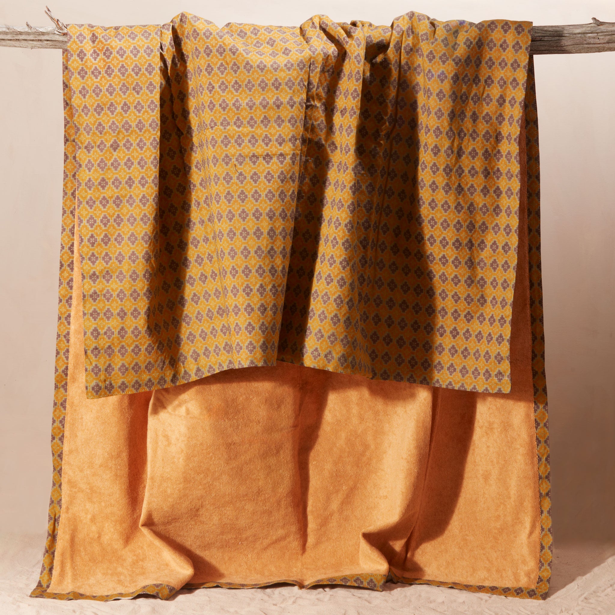 A draped kanga towel in warm mustard yellow and brown, featuring a patterned cotton side and a yellow-orange terry cloth side, hanging from a rustic wooden branch against a neutral beige background.