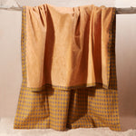 The yellow-orange terry cloth side of a kanga towel in warm mustard yellow and brown, featuring a patterned cotton side, hanging from a rustic wooden branch against a neutral beige background.