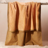 The yellow-orange terry cloth side of a kanga towel in warm mustard yellow and brown, featuring a patterned cotton side, hanging from a rustic wooden branch against a neutral beige background.