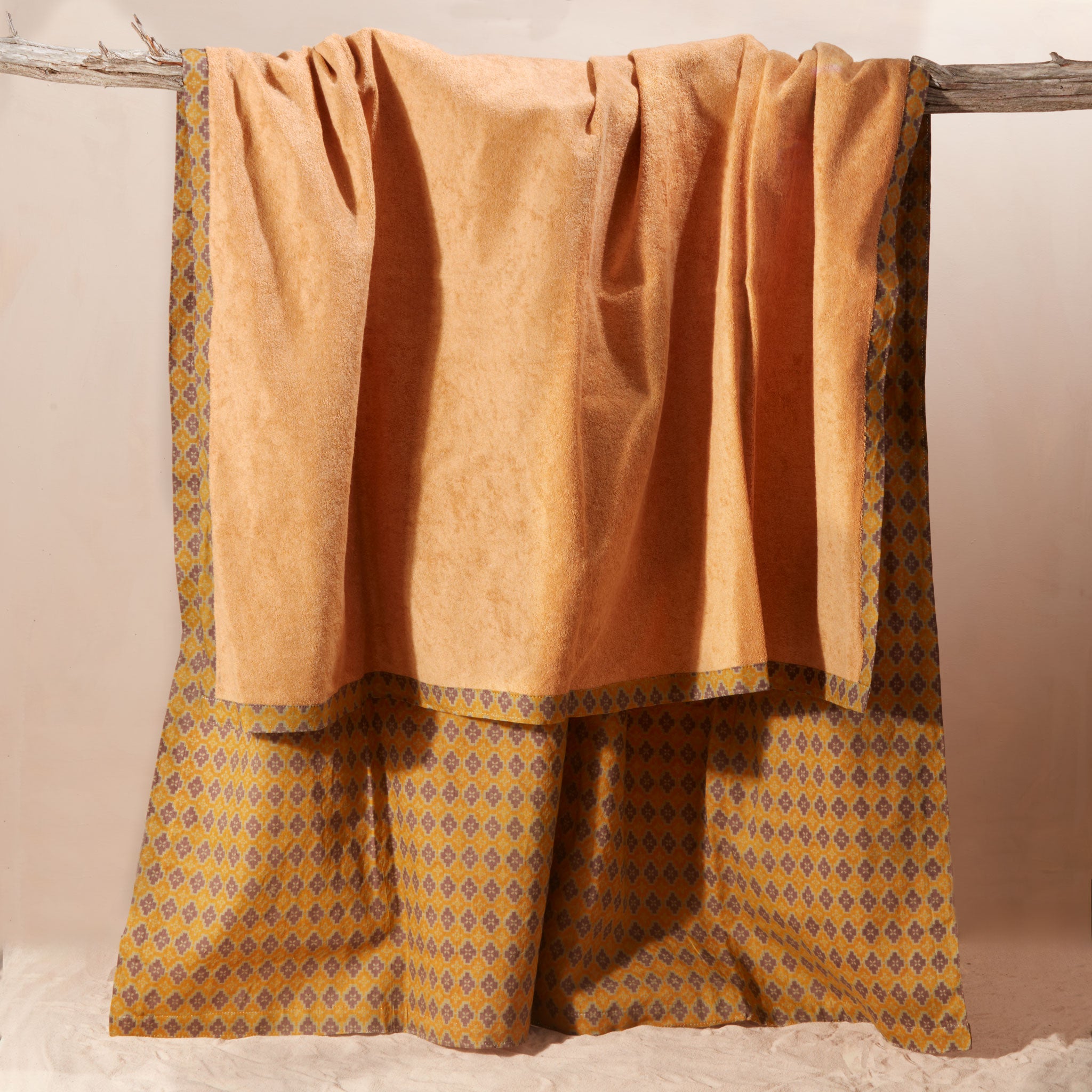 The yellow-orange terry cloth side of a kanga towel in warm mustard yellow and brown, featuring a patterned cotton side, hanging from a rustic wooden branch against a neutral beige background.