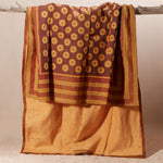 A draped kanga towel of rust-brown and mustard-yellow fabric with circular motifs and striped borders with a yellow-orange terry cloth lining, hanging from a rustic branch against a plain beige background.