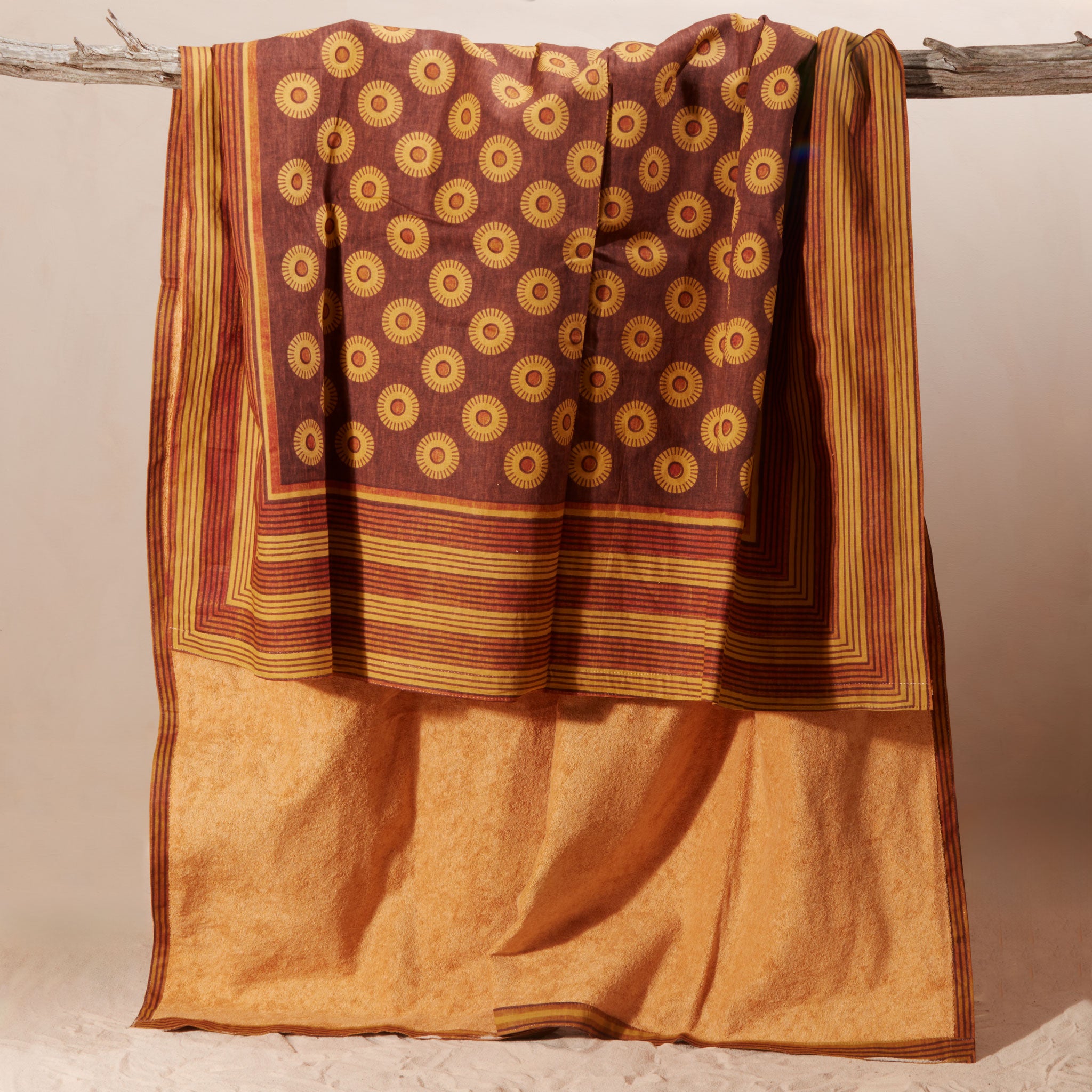 A draped kanga towel of rust-brown and mustard-yellow fabric with circular motifs and striped borders with a yellow-orange terry cloth lining, hanging from a rustic branch against a plain beige background.