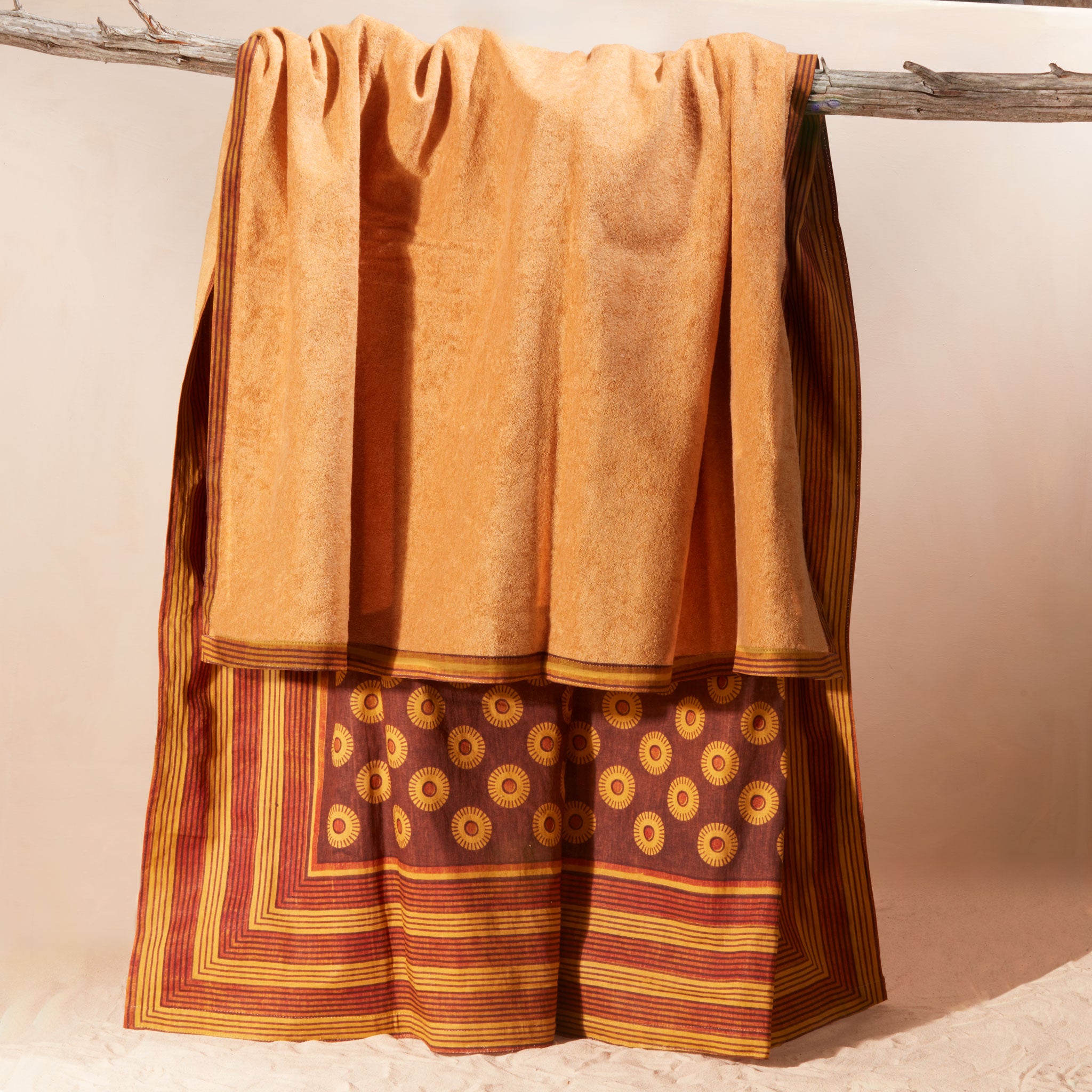 The yellow-orange terry cloth lining of a  draped kanga towel of rust-brown and mustard-yellow fabric with circular motifs and striped borders, hanging from a rustic branch against a plain beige background.